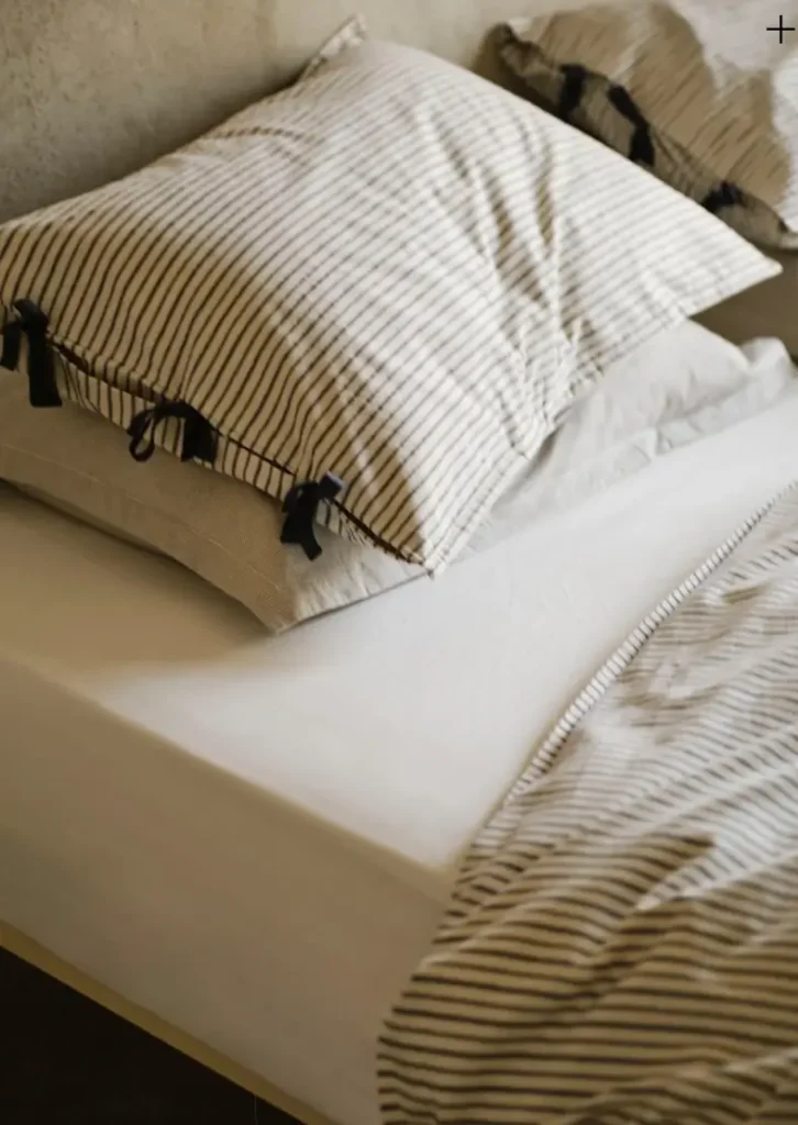 Cozy bed styled with Rowan Print Bedding, showing soft beige and black fine-striped pillowcases with elegant black tie closures on the envelope opening. The pillows rest on crisp white sheets alongside a matching striped duvet, creating a calm, timeless, and textured neutral bedroom scene