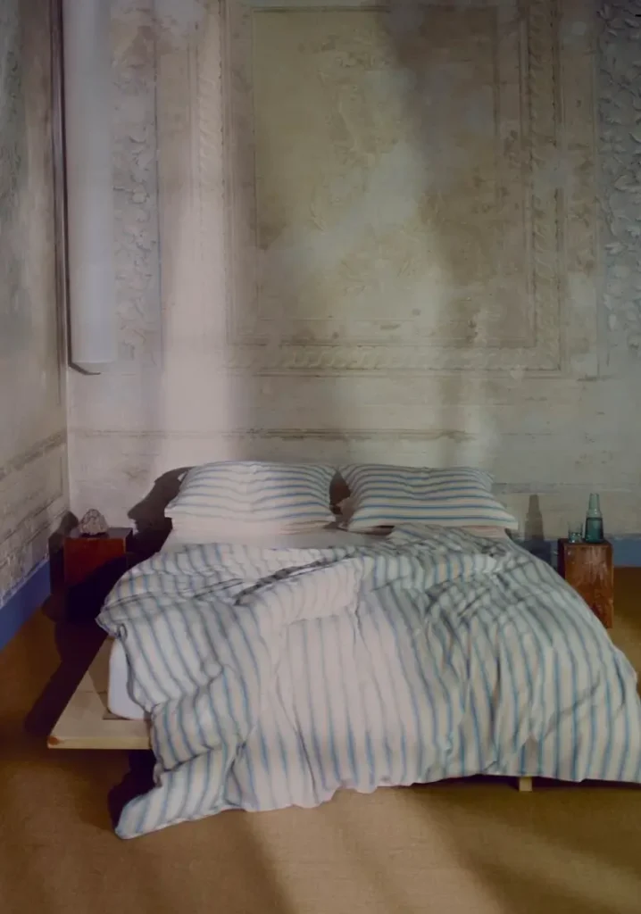 Serene bedroom featuring Rowan Print Bedding in soft blue and white stripes. A low platform bed is dressed with a relaxed striped duvet and matching pillows against textured, aged plaster walls. Natural light casts gentle shadows across the minimalist space, creating a calm and timeless atmosphere