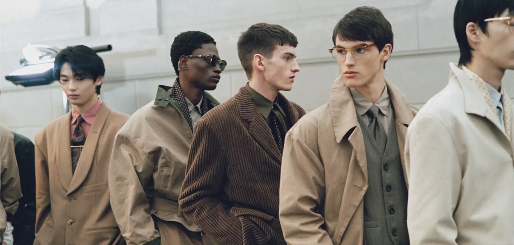 A line of men stand side by side in a softly lit interior, dressed in coordinated, earth-toned tailoring that blends structured suits with relaxed outerwear. Layers of trench coats, textured jackets, and shirts with ties create a cohesive, refined palette of beige, olive, and brown. Subtle variations—like corduroy textures, oversized silhouettes, and accessories such as glasses and sunglasses—add individuality, while their composed expressions and forward-facing stances evoke a calm, editorial runway atmosphere