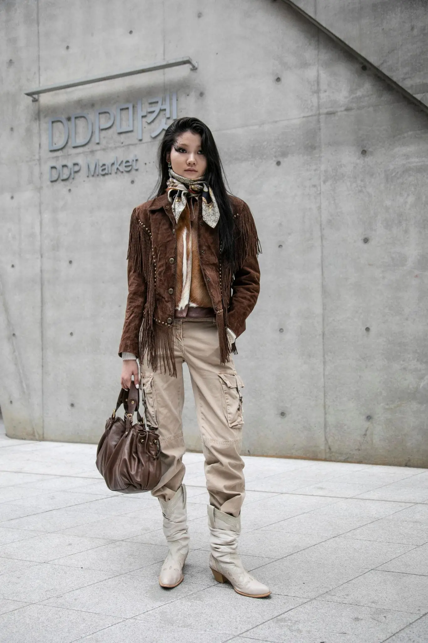 Street-style portrait of a woman in a brown fringed suede jacket, patterned silk scarf, and beige cargo pants, paired with slouchy off-white heeled boots, standing against a minimalist concrete wall while holding a large brown leather handbag