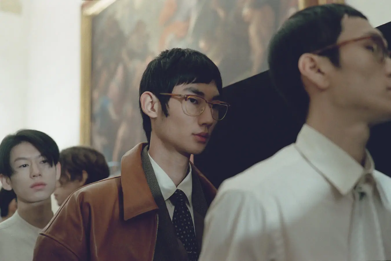 A group of young men move in line through a softly lit interior, dressed in refined, layered tailoring with a muted, classic palette. The central figure wears thin-framed glasses, a brown leather jacket over a shirt and patterned tie, blending casual and formal elements. Others around him appear in crisp shirts, ties, and understated outerwear, their calm expressions and forward motion creating a composed, quietly cinematic scene reminiscent of a backstage or runway moment