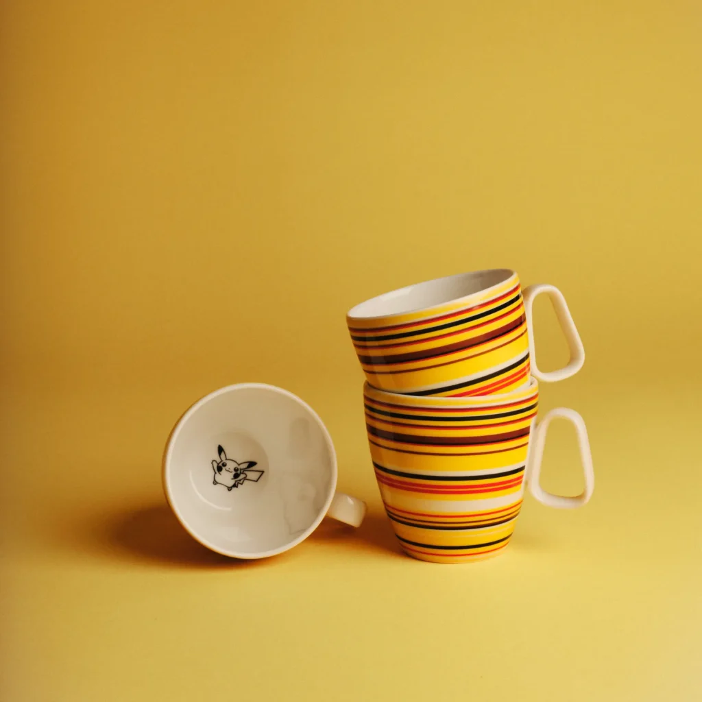 A trio of ceramic mugs arranged against a warm yellow background, each decorated with bold horizontal stripes in yellow, orange, red, and black. Two mugs are stacked upright, slightly angled, while a third lies on its side, revealing a small black-and-white Pikachu illustration inside. The composition highlights the vibrant striping and playful interior detail with a clean, minimal presentation