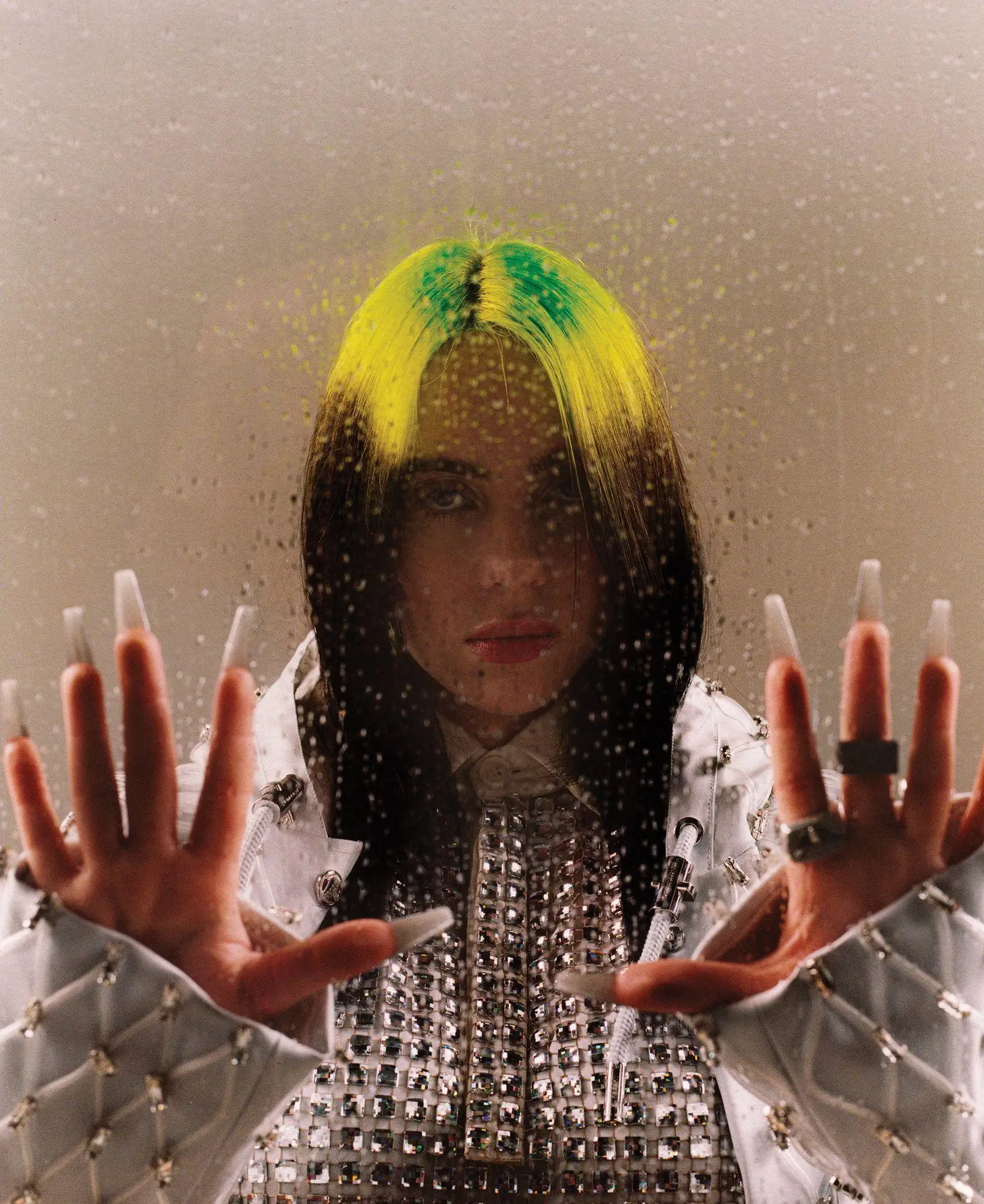 Portrait of Billie Eilish behind a rain-speckled glass surface, her hands pressed toward the viewer. She wears a textured white jacket with metallic embellishments, and her dark hair features bright neon green roots. Soft, warm lighting highlights her calm, direct expression against a neutral background
