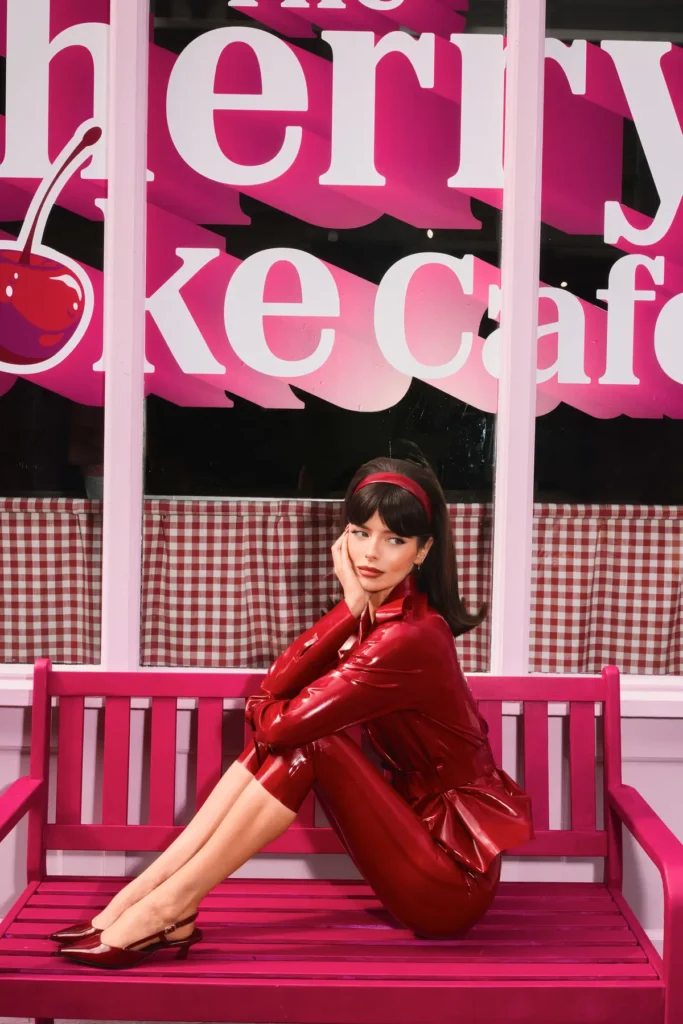 A polished, almost cinematic portrait situates the subject on a vivid pink bench, framed by a café-style window emblazoned with oversized “Cherry Coke Café” lettering. The composition feels deliberately symmetrical and controlled, with the bold typography looming large behind her—part backdrop, part statement. She sits in a compact, sculptural pose—knees drawn in, arms wrapped lightly, chin resting against her hand—creating a sense of poised stillness. The glossy red outfit, sharply tailored and reflective, catches the light in clean highlights, reinforcing the high-fashion, campaign-driven tone. A matching red headband ties the look together, echoing both the product palette and a retro, mid-century femininity. The environment layers texture and nostalgia: gingham café curtains line the window, while the pink-painted bench and trim push the space into a stylized, almost dollhouse-like interpretation of a classic soda shop. The color story—deep reds, bubblegum pinks, and soft whites—remains tightly cohesive throughout. Lighting is bright and frontal, flattening shadows just enough to emphasize color and surface over depth, giving the image a crisp, editorial finish. Unlike more candid scenes, this moment is fully composed—every element aligned for visual impact. Overall, the image reads as a hero vignette within the broader installation: fashion-forward, brand-saturated, and intentionally iconic, where subject, set, and signage merge into a single, highly controlled visual identity