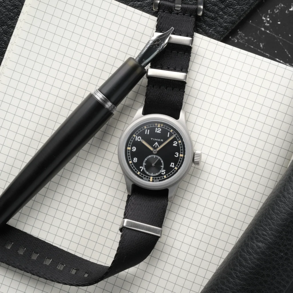 A classic field watch with a black dial and black fabric strap rests on a grid-lined notebook, paired with a black fountain pen and leather accessories, highlighting a refined, utilitarian desk setup