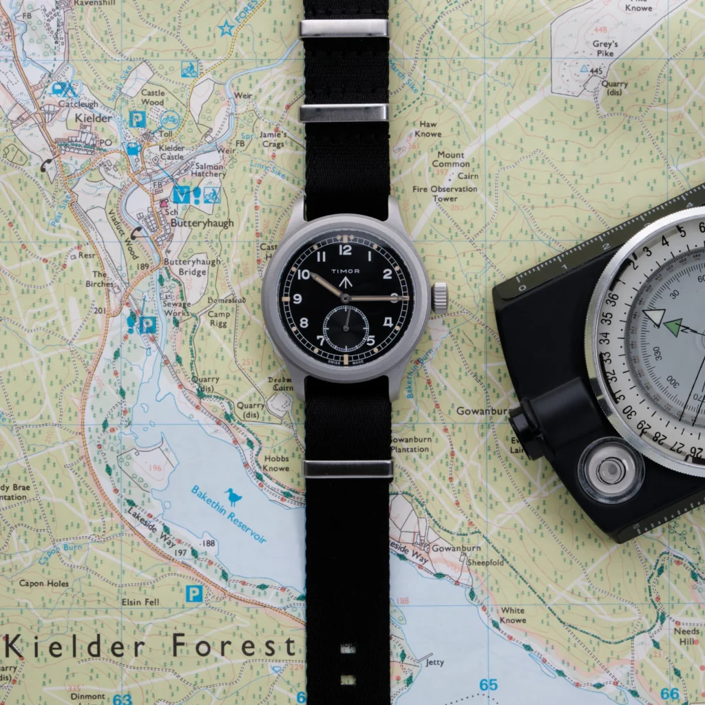 A classic field watch with a black dial and black fabric strap is positioned over a detailed topographic map, accompanied by a navigation tool, emphasizing an outdoor exploration and navigation theme
