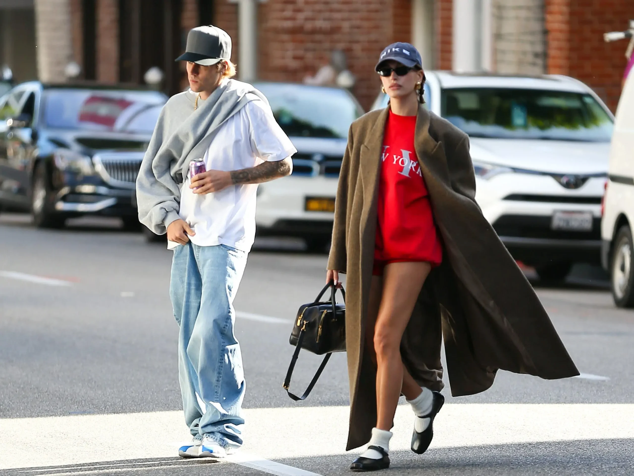 Justin Bieber and Hailey Bieber walking together on a city street in casual daytime outfits. Justin wears an oversized white t-shirt, light-wash baggy jeans, a grey baseball cap, and a grey sweatshirt draped over his shoulders while holding a drink. Hailey wears a bright red 'New York' sweatshirt, a long brown overcoat, black Mary Jane shoes with white socks, a navy cap, and carries a black handbag