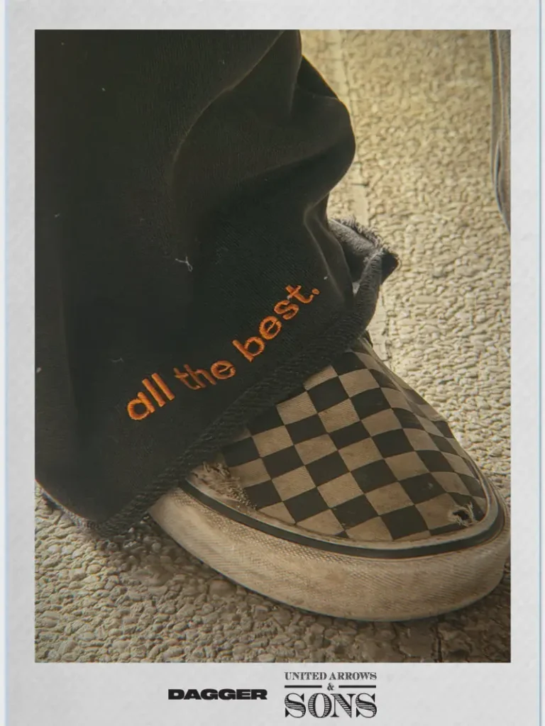 DAGGER x United Arrows & Sons campaign close-up featuring frayed black sweatpants embroidered with “all the best” in orange, paired with worn checkerboard slip-on sneakers, captured in a textured, vintage-toned streetwear aesthetic