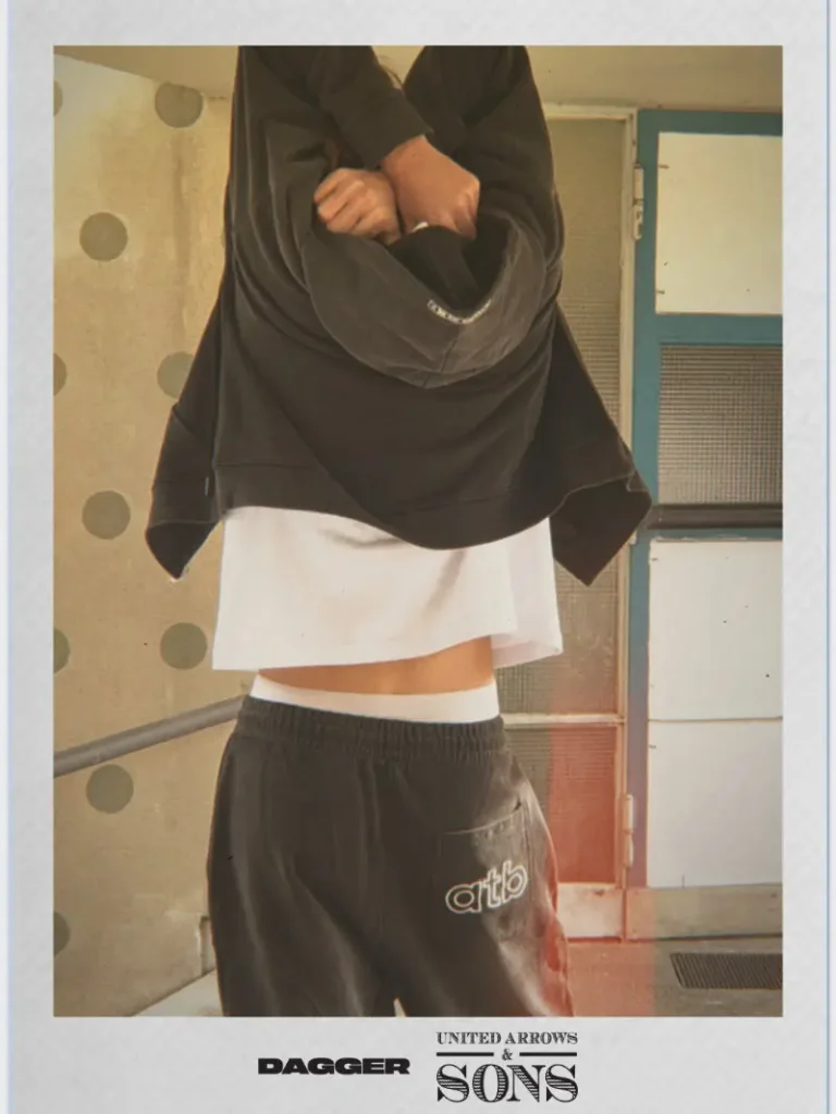 DAGGER x United Arrows & Sons campaign image showing a model lifting a black hoodie to reveal layered styling with a white tee and relaxed sweatpants, captured in warm natural light with a raw, candid streetwear feel