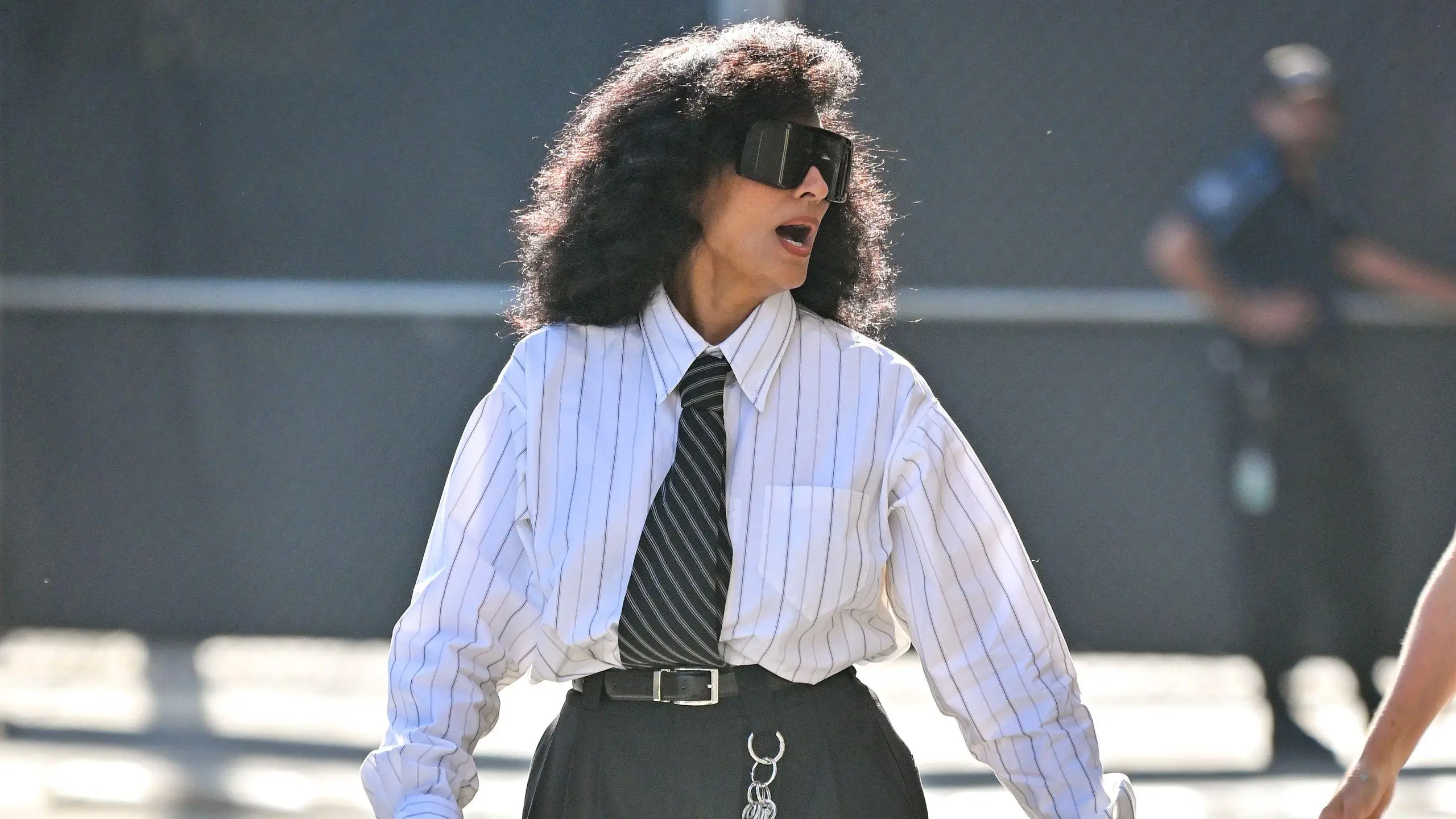 Tracee Ellis with voluminous curly hair wearing oversized black visor-style sunglasses, a loose white pinstriped button-down shirt, and a striped tie tucked into high-waisted black trousers with a belt and chain detail, captured mid-movement outdoors in bright sunlight with a blurred background
