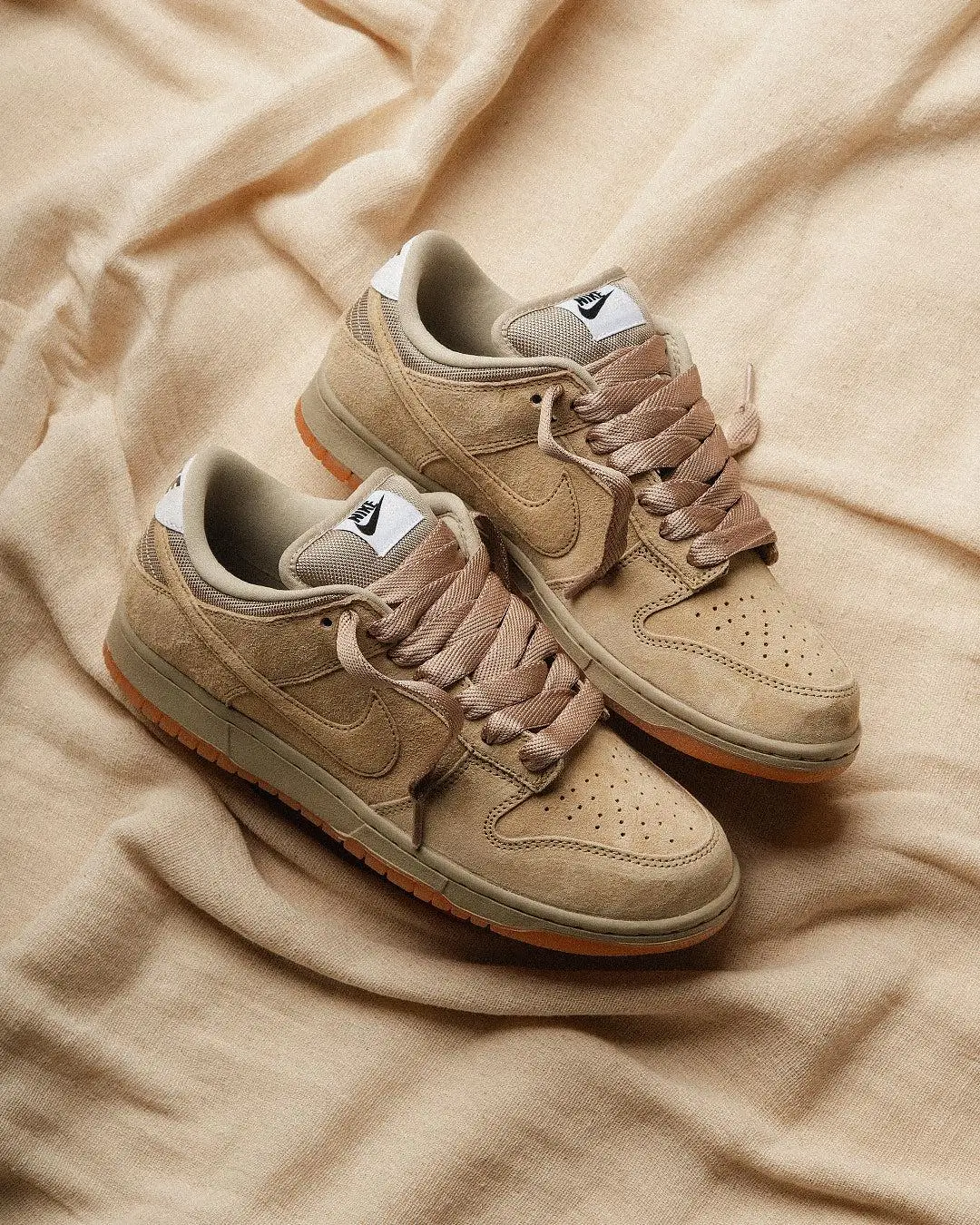 Nike SB Dunk Low Pro in Vachetta Tan and Parachute Beige, shown as a pair on softly draped beige fabric, featuring a monochromatic suede and canvas upper, tonal laces, subtle Swoosh branding, and a warm gum outsole view along the edges