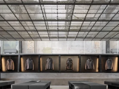 A stark, industrial retail interior defined by symmetry and restraint. A translucent grid ceiling spans overhead, revealing exposed truss structures and diffused daylight. Along the back wall, a continuous row of recessed display boxes showcases technical outerwear jackets, each individually lit to emphasize material and form. In the foreground, low, modular black seating blocks are arranged with geometric precision, reinforcing the gallery-like atmosphere. Raw concrete surfaces, metal mesh detailing, and a muted tonal palette create a space that feels both utilitarian and curated, where architecture and product are presented with equal emphasis
