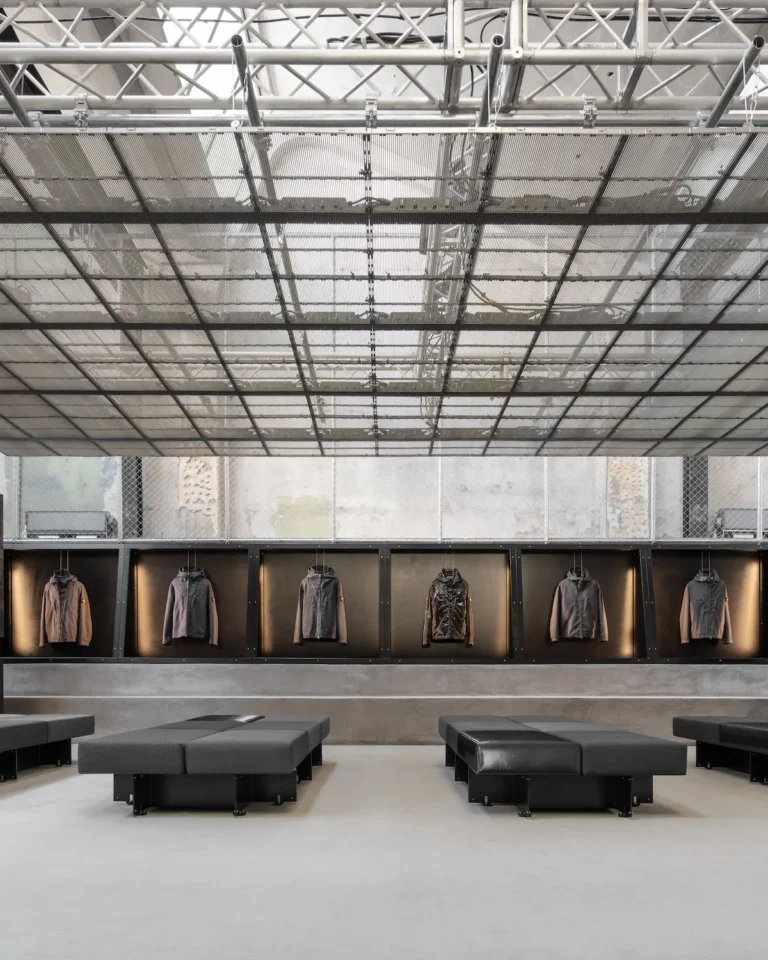 A stark, industrial retail interior defined by symmetry and restraint. A translucent grid ceiling spans overhead, revealing exposed truss structures and diffused daylight. Along the back wall, a continuous row of recessed display boxes showcases technical outerwear jackets, each individually lit to emphasize material and form. In the foreground, low, modular black seating blocks are arranged with geometric precision, reinforcing the gallery-like atmosphere. Raw concrete surfaces, metal mesh detailing, and a muted tonal palette create a space that feels both utilitarian and curated, where architecture and product are presented with equal emphasis
