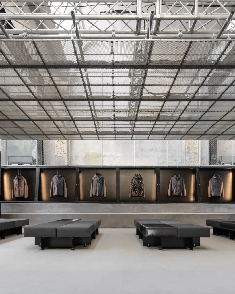 A stark, industrial retail interior defined by symmetry and restraint. A translucent grid ceiling spans overhead, revealing exposed truss structures and diffused daylight. Along the back wall, a continuous row of recessed display boxes showcases technical outerwear jackets, each individually lit to emphasize material and form. In the foreground, low, modular black seating blocks are arranged with geometric precision, reinforcing the gallery-like atmosphere. Raw concrete surfaces, metal mesh detailing, and a muted tonal palette create a space that feels both utilitarian and curated, where architecture and product are presented with equal emphasis