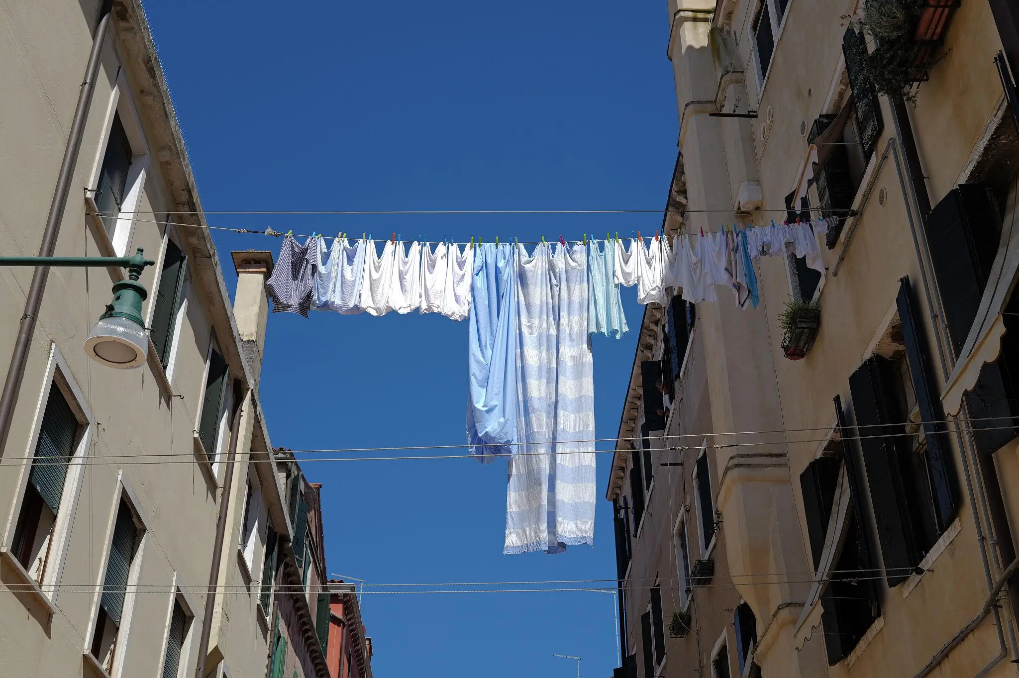 Laundry in white and light blue fabrics hanging on a clothesline stretched between two buildings, set against a clear blue sky in a narrow street scene