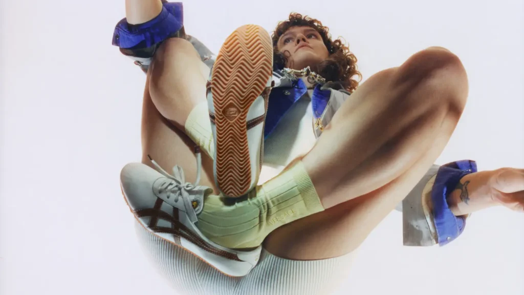 ow-angle campaign image of a model wearing Onitsuka Tiger x Versace Tai-Chi Sakura shoe, highlighting the gum outsole tread, metallic leather panels, and streamlined low-profile silhouette against a bright minimal backdrop