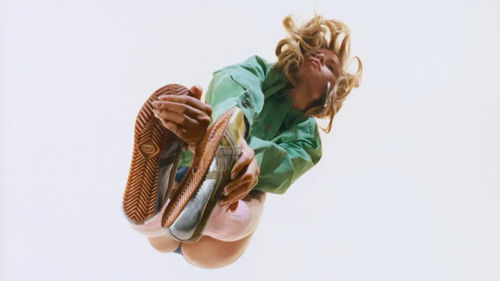Low-angle campaign image of a model in a green jacket wearing Onitsuka Tiger x Versace Tai-Chi Sakura shoe, highlighting the gum outsole tread, metallic leather upper, and elongated minimal silhouette against a clear sky backdrop