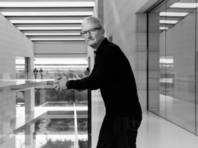 Tim Cook stands in a minimalist, glass-lined corridor at Apple Park, leaning on a railing in a composed black-and-white portrait, his posture calm and reflective against the clean architectural lines