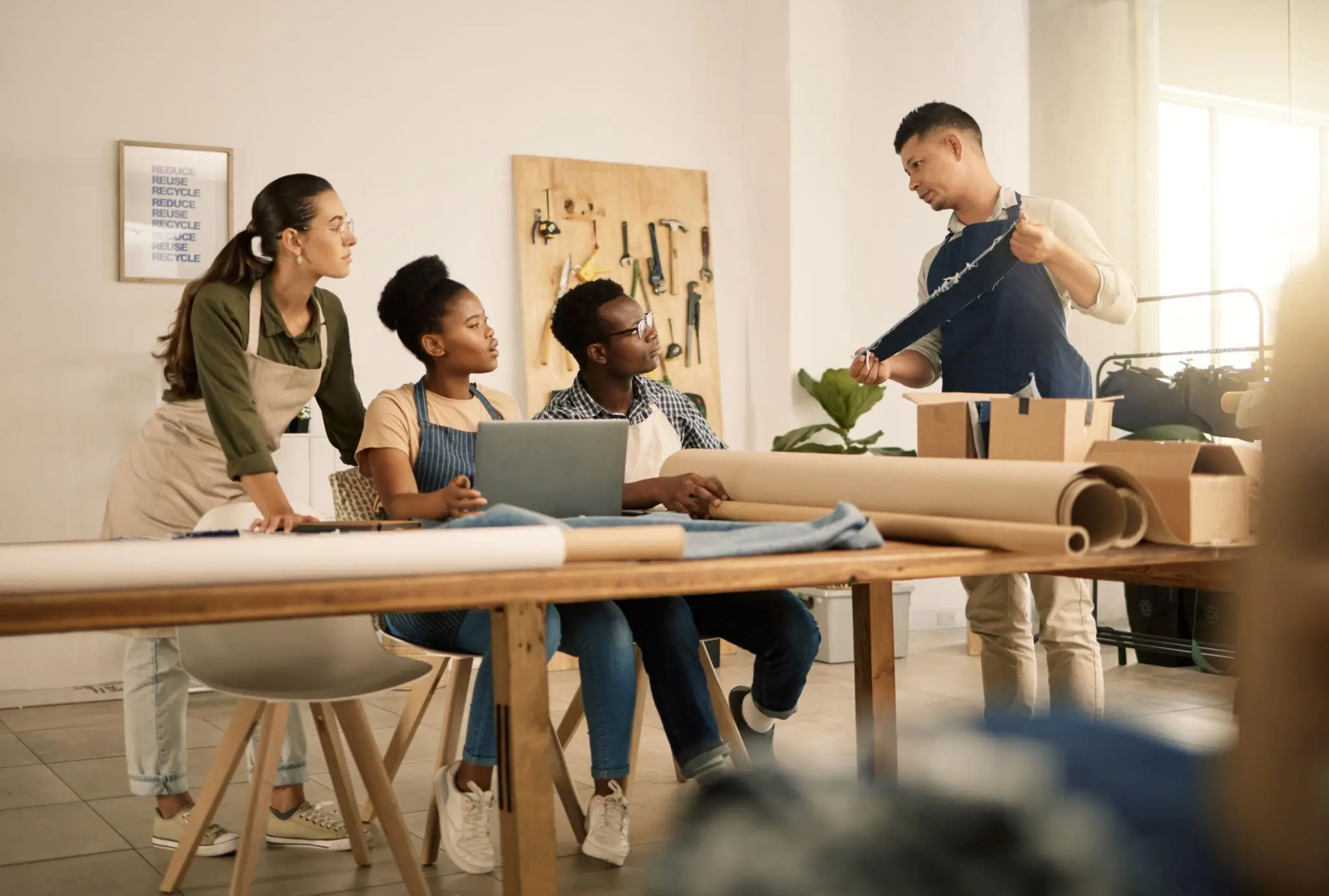 A small group of people gather around a wooden worktable in a bright, workshop-style space, listening as one person in an apron holds up a piece of fabric or material to demonstrate. Others sit or lean in attentively, with a laptop open on the table alongside rolls of paper and textiles. Tools hang neatly on the wall behind, and soft daylight from nearby windows creates a warm, collaborative, hands-on learning atmosphere