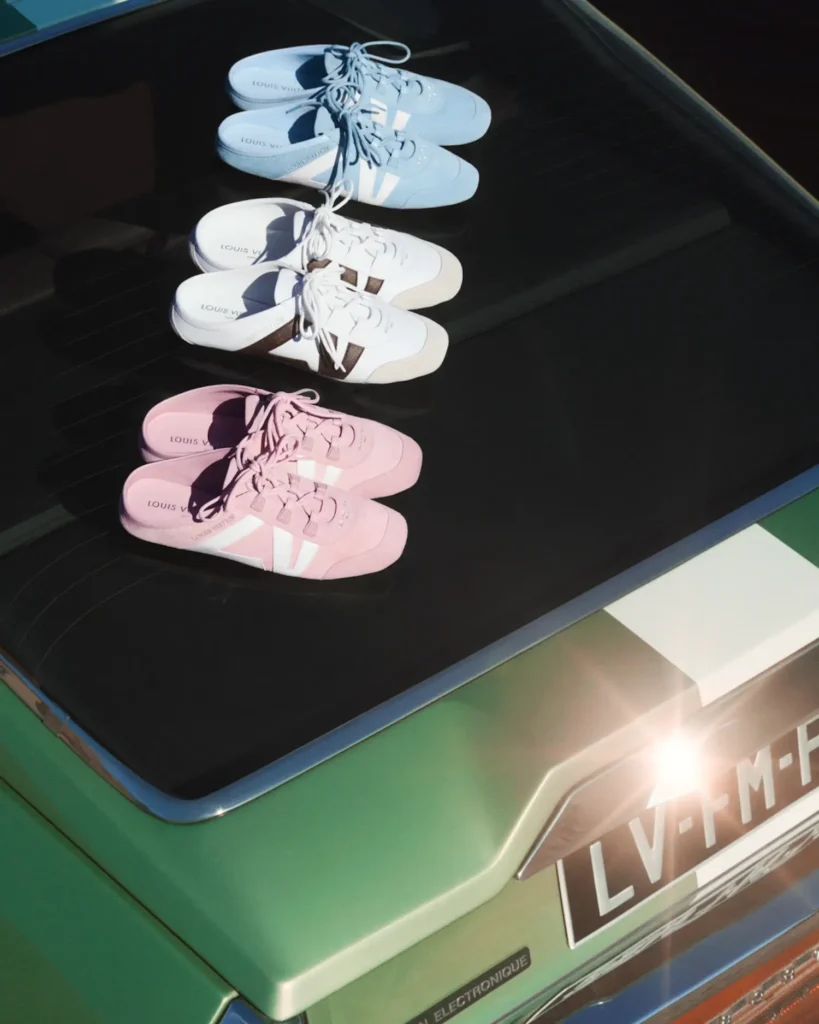 Three pairs of Louis Vuitton mule sneakers are neatly arranged on the rear window of a green vintage car, displayed in soft pastel blue, white with brown accents, and pink. The shoes are laced and aligned in a clean row, their branding subtly visible on the insoles. Sunlight reflects off the glass and metallic paint, creating a polished, editorial-style presentation that emphasizes color variation and design detail
