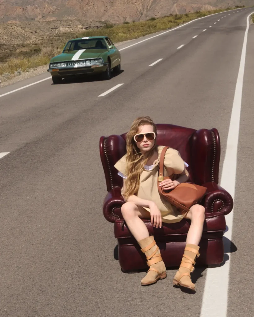 A young woman sits relaxed in a deep burgundy leather armchair placed directly on an open desert highway, her legs stretched forward with an effortless, nonchalant posture. She wears oversized sunglasses, a loose beige outfit, strappy tan boots, and carries a structured brown shoulder bag. In the distance behind her, a green vintage car approaches along the empty road, while sunlit mountains and dry terrain frame the scene, creating a surreal, editorial juxtaposition of comfort and motion