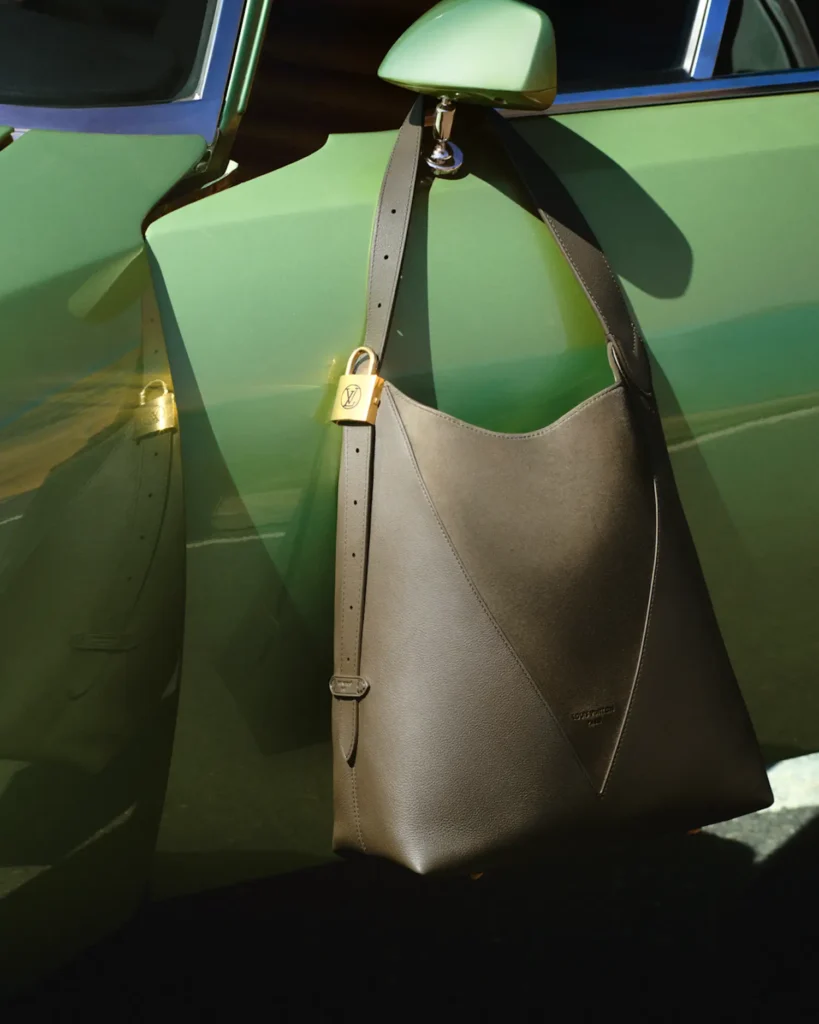 A structured brown leather shoulder bag hangs from the side mirror of a green vintage car, its long strap draped neatly with a gold padlock detail adding a refined accent. The smooth, sculptural shape of the bag contrasts with the glossy car surface, where sunlight casts sharp reflections and shadows, emphasizing texture, craftsmanship, and a clean, haute aesthetic