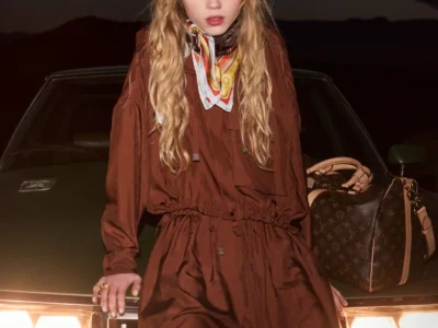 A young woman stands against the front of a vintage car at night, illuminated by its glowing headlights. She wears a loose brown jumpsuit paired with a patterned silk scarf tied at the neck, her wavy hair falling naturally around her face. A structured Louis Vuitton monogram bag rests beside her on the hood, while the dark desert horizon behind her enhances the moody, cinematic atmosphere