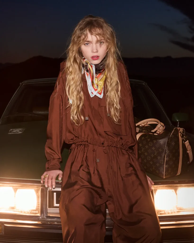 A young woman stands against the front of a vintage car at night, illuminated by its glowing headlights. She wears a loose brown jumpsuit paired with a patterned silk scarf tied at the neck, her wavy hair falling naturally around her face. A structured Louis Vuitton monogram bag rests beside her on the hood, while the dark desert horizon behind her enhances the moody, cinematic atmosphere