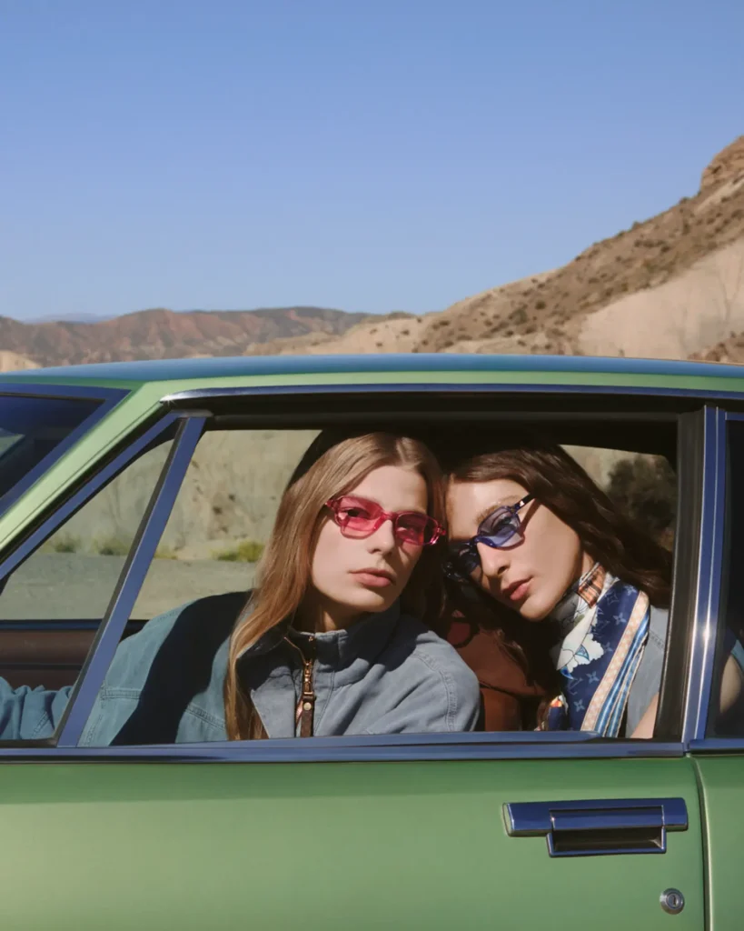Two young women sit side by side inside a green vintage car, leaning slightly toward each other as they look out through the open window. Both wear tinted sunglasses—one in soft red, the other in cool blue—paired with relaxed, fashion-forward outfits and a patterned silk scarf. Sunlight illuminates their faces against a backdrop of pale desert hills, creating a crisp, modern editorial moment with a subtle sense of intimacy and ease