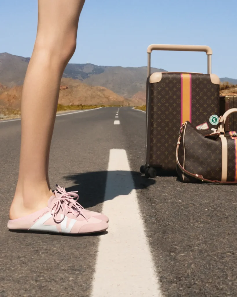A close-up of a woman’s legs standing on the center line of an open desert road, wearing soft pink Louis Vuitton mule sneakers with laces. Beside her, a monogram rolling suitcase with a colorful stripe and a matching duffel bag rest on the asphalt, ready for travel. The long, empty road stretches toward distant mountains under a clear sky, creating a clean, directional composition that emphasizes movement, style, and journey
