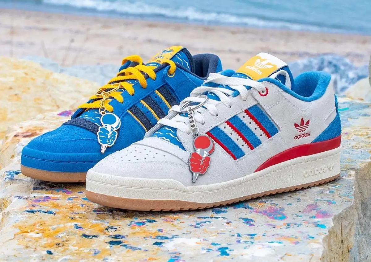 Pair of Adidas shoes in blue and white colorways with red and yellow accents, featuring Three Stripes branding, gum soles, and playful keychain charms, displayed on a paint-splattered surface by the seaside