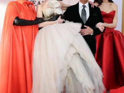 A group of four people pose together on a red carpet, standing closely with arms linked in a warm, celebratory moment. They are dressed in formal eveningwear, suggesting a film premiere or major event. On the left, a woman wears a bold, flowing red-orange cape-style gown paired with long black gloves and oversized sunglasses, projecting a dramatic, high-fashion presence. Next to her, a woman in an elaborate gown combines a sculpted, metallic-toned bodice with a voluminous, layered tulle skirt in soft off-white, creating a striking contrast of structure and softness; her expression is lively and joyful