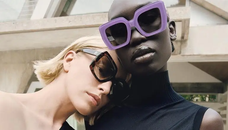 Two models posing closely outdoors, one with short blonde hair wearing oversized brown-tinted square sunglasses, the other with a shaved head wearing bold purple geometric sunglasses and a black sleeveless top, set against a modern architectural backdrop