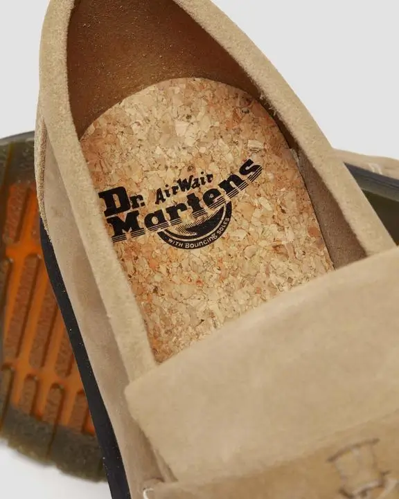 A close-up of a beige suede slip-on loafer highlighting its cork-textured footbed, stamped with the Dr. Martens AirWair logo. The soft suede upper frames the interior, while the translucent brown outsole is partially visible beneath, emphasizing a comfort-focused, casual construction with signature branding