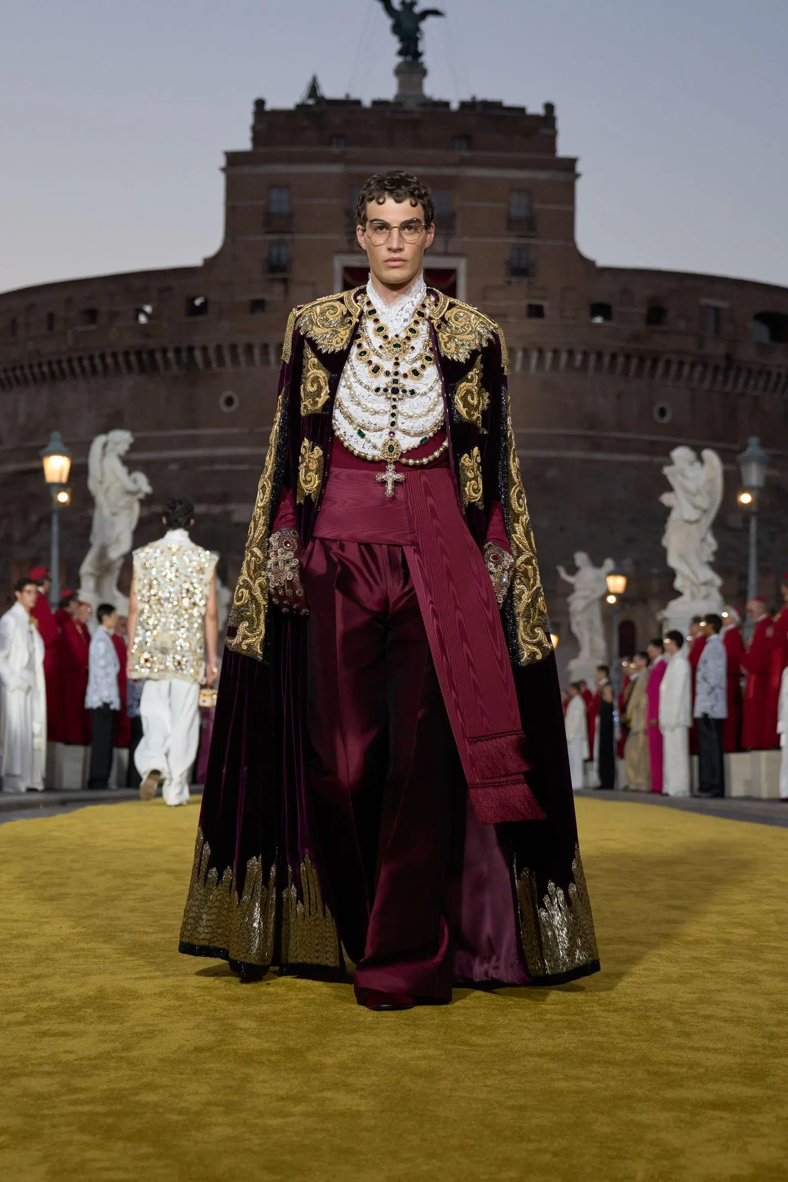 Model walking a runway in an ornate burgundy and gold ensemble, featuring a richly embroidered cape, layered statement jewelry with crosses and gemstones, and wide-leg trousers, set against a historic architectural backdrop with statues and an audience lining the scene