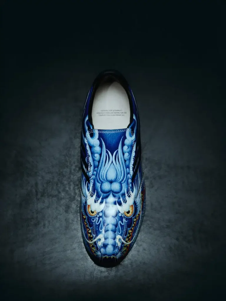 Top-down view of a blue Y-3 F50 TUNIT shoe featuring a detailed dragon graphic across the upper, set against a dark textured background