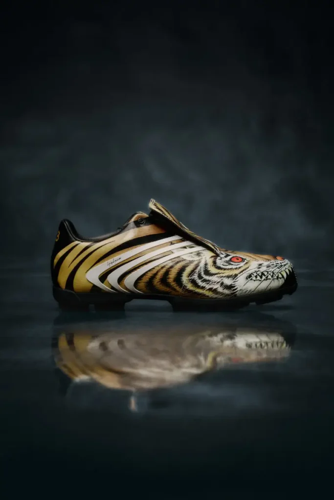 Side profile of a yellow Y-3 F50 TUNIT football boot with tiger-inspired stripes and red eye detailing, featuring a fold-over tongue and white adidas stripes on a dark reflective studio surface