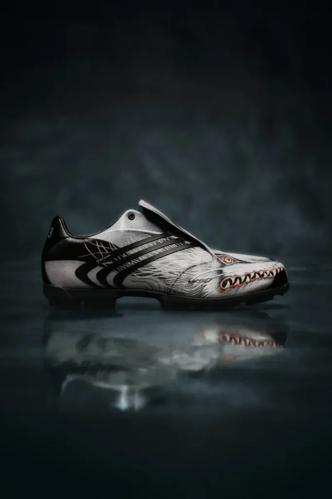 Side profile of a black and silver Y-3 F50 TUNIT football boot with wolf-inspired graphics, fold-over tongue, and black adidas stripes on a dark reflective studio surface