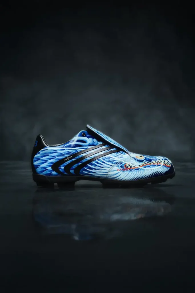 Side profile of a blue Y-3 F50 TUNIT football boot with dragon-scale graphics, fold-over tongue, and black adidas stripes on a dark studio background