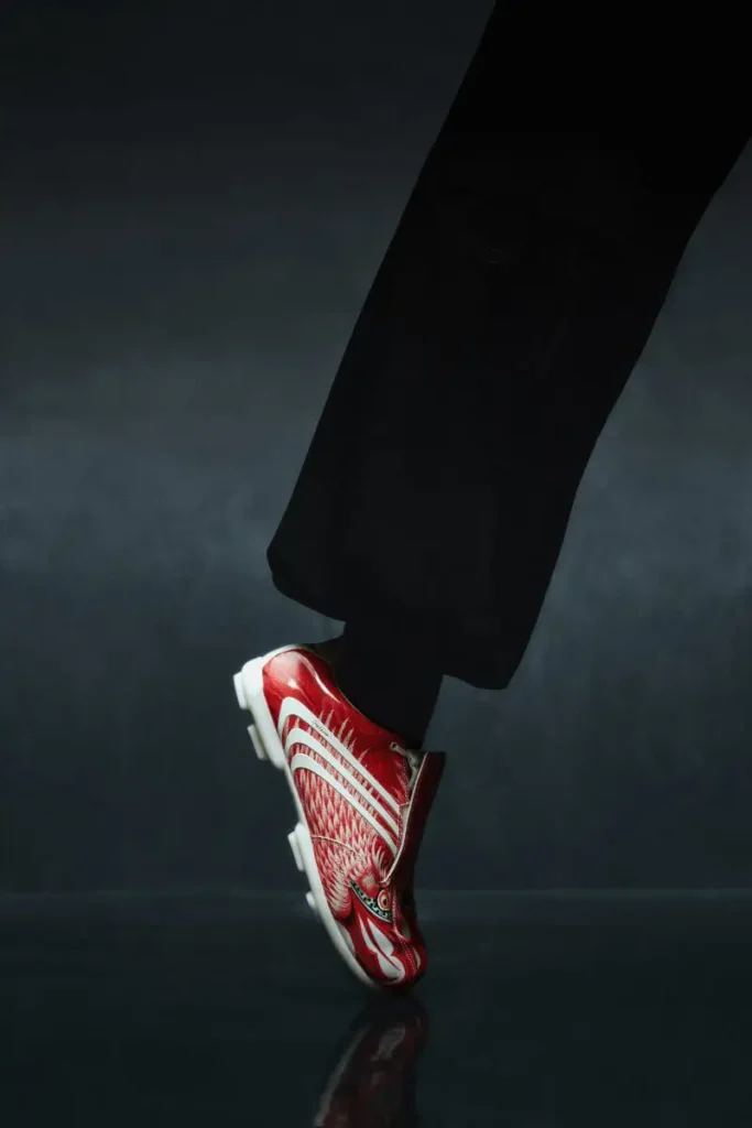 Red Y-3 F50 TUNIT football boot with eagle-inspired graphic worn with black trousers, captured mid-step against a dark studio backdrop