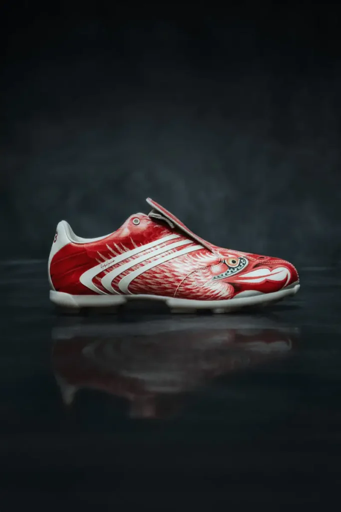 Side profile of a red Y-3 F50 TUNIT football boot with eagle-inspired graphics, fold-over tongue, and white adidas stripes on a dark studio background