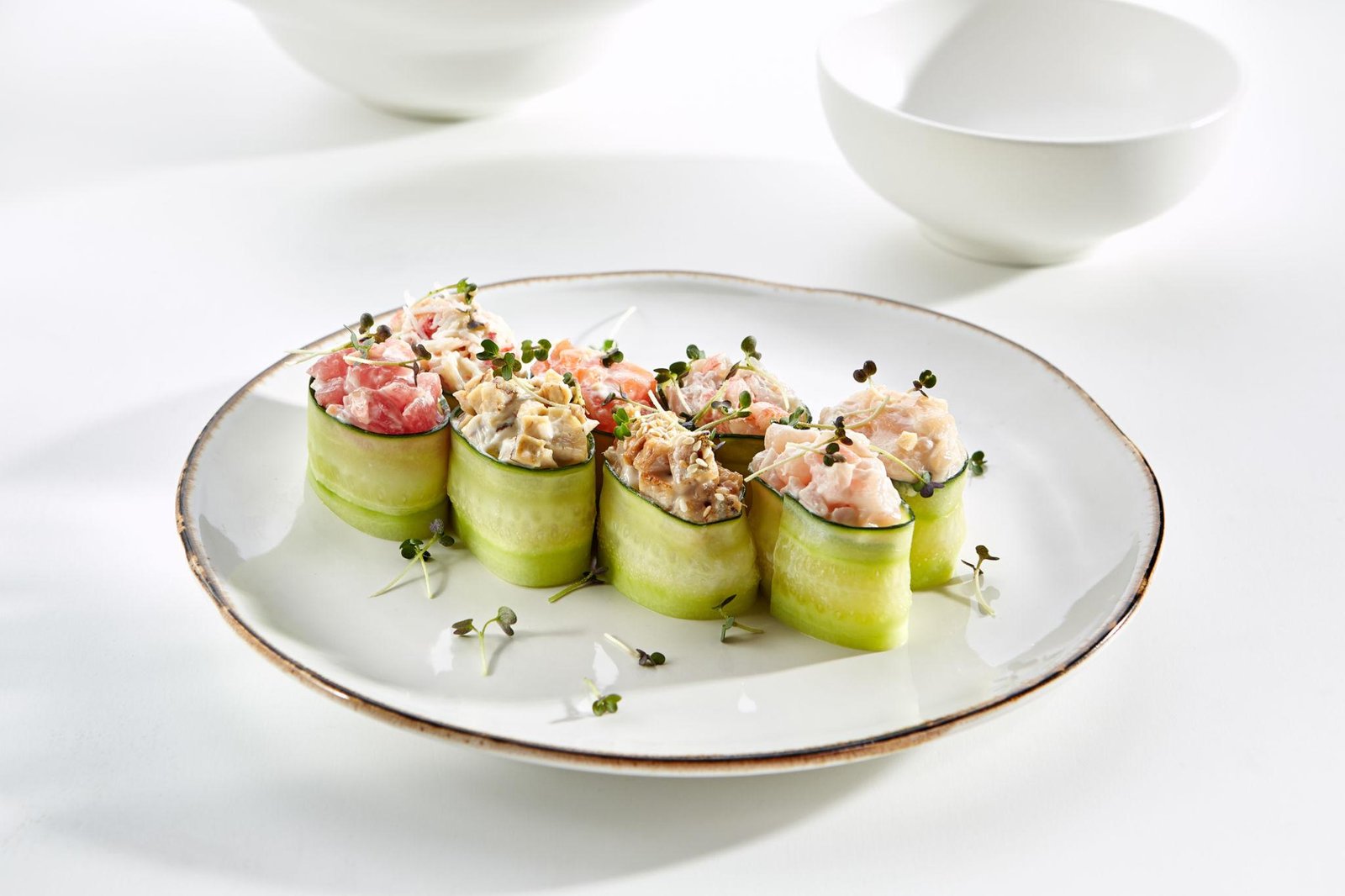 Minimalist plated appetizer featuring neat rolls of thinly sliced cucumber filled with a mix of seafood and creamy toppings, garnished with microgreens and arranged on a white ceramic plate against a clean, bright table setting