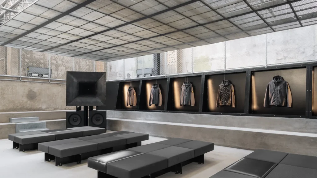 A refined industrial showroom interior where architecture, sound, and apparel converge. Modular black seating blocks are arranged in clean, linear rows, facing a large sculptural horn speaker mounted above dual subwoofers, anchoring the space as a focal installation. Overhead, a translucent grid ceiling diffuses natural light while revealing exposed truss structures, reinforcing the raw, engineered aesthetic. Along the right wall, a sequence of recessed display compartments presents technical jackets, each individually spotlighted to highlight texture and construction. The backdrop of unfinished concrete and mesh paneling adds a utilitarian edge, while the careful spacing and lighting elevate the garments to gallery-like objects. The overall composition feels deliberate and immersive—part listening room, part fashion exhibition—where sound, structure, and product are given equal weight