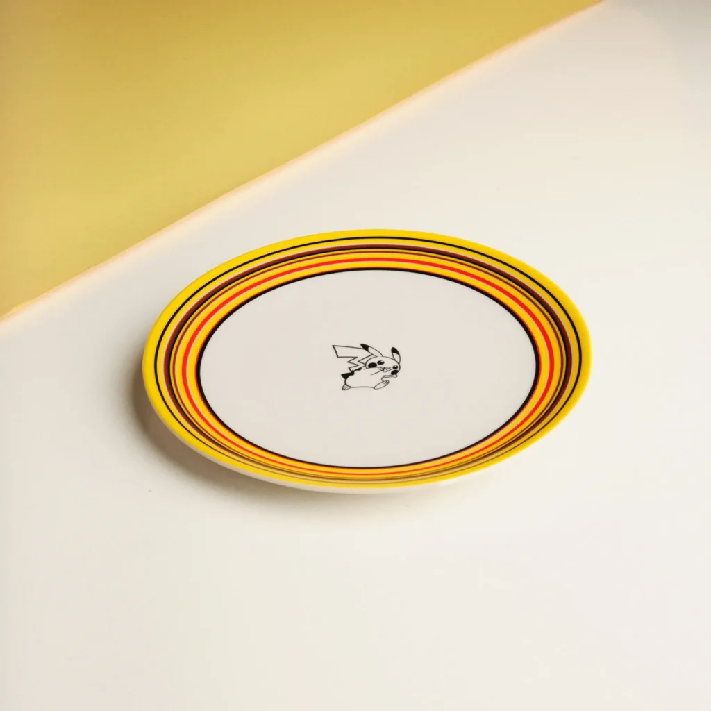 A single ceramic plate placed on a clean, two-tone surface of soft white and warm yellow, featuring concentric rings in yellow, orange, red, and black around the rim. At the center, a small black-and-white Pikachu illustration adds a play focal point. The minimal composition emphasizes the bold striping and refined, modern presentation