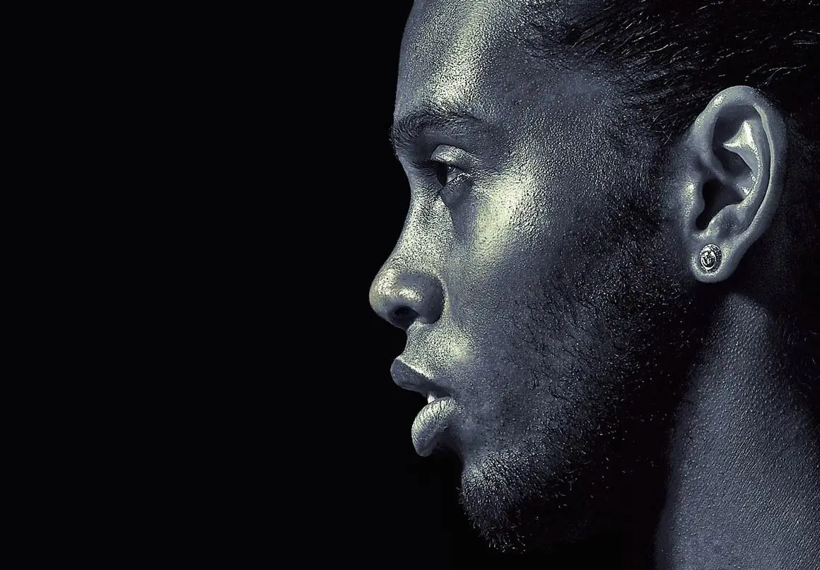 High-contrast side profile portrait of Ronladinho with textured skin and a subtle metallic sheen, wearing a stud earring against a deep black background, lit dramatically to emphasize facial contours
