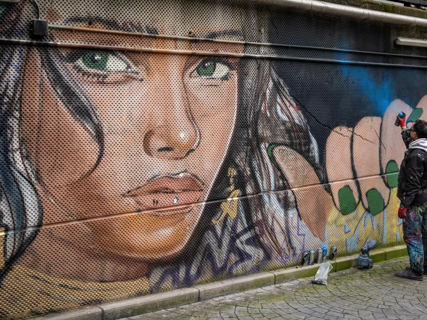 A street artist stands beside a large perforated metal wall, actively spray-painting a vibrant mural that features a close-up portrait with striking green eyes. Spray cans and tools are scattered along the ground, while the textured surface and bold colors create a dynamic, urban scene that captures the process of graffiti art in progress