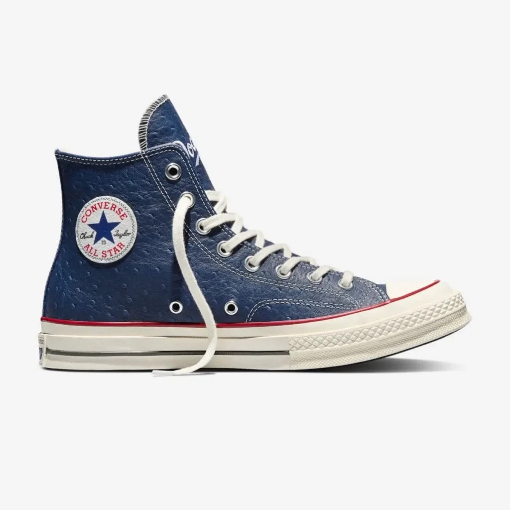 Side view of a navy high-top shoe with white laces and cream rubber sole, featuring a textured upper, red piping detail, and the Converse All Star circular patch on the ankle