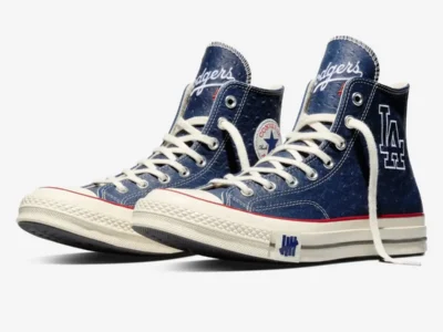 Angled view of a pair of navy high-top shoe with white laces and cream rubber soles, featuring textured leather uppers, red stripe detailing, “Dodgers” script on the tongues, an “LA” logo on one shoe, Converse All Star patch on the other, and UNDEFEATED tally branding near the midsole