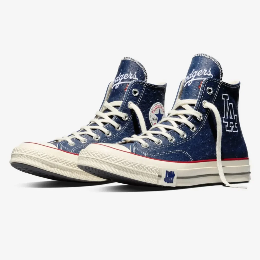 Angled view of a pair of navy high-top shoe with white laces and cream rubber soles, featuring textured leather uppers, red stripe detailing, “Dodgers” script on the tongues, an “LA” logo on one shoe, Converse All Star patch on the other, and UNDEFEATED tally branding near the midsole