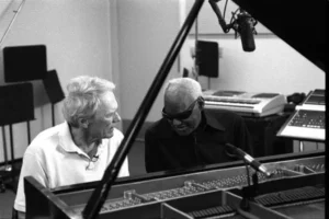 Clint Eastwood seated beside a collaborator at a grand piano in a recording studio, both leaning in conversation under suspended microphones, captured in black and white to emphasize a quiet, intimate creative exchange